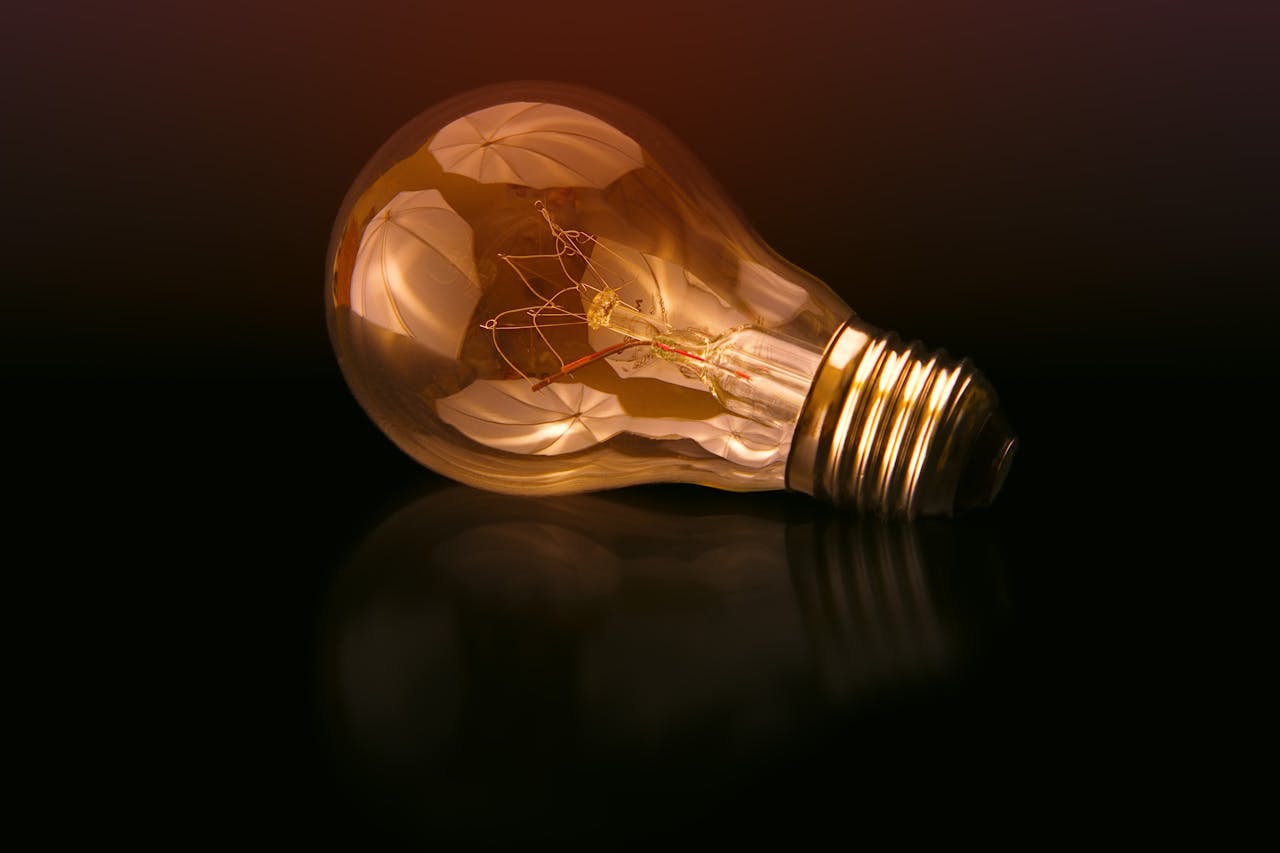 who-we-are A detailed close-up of a light bulb showcasing its intricate filament and glowing glass against a dark reflective surface.